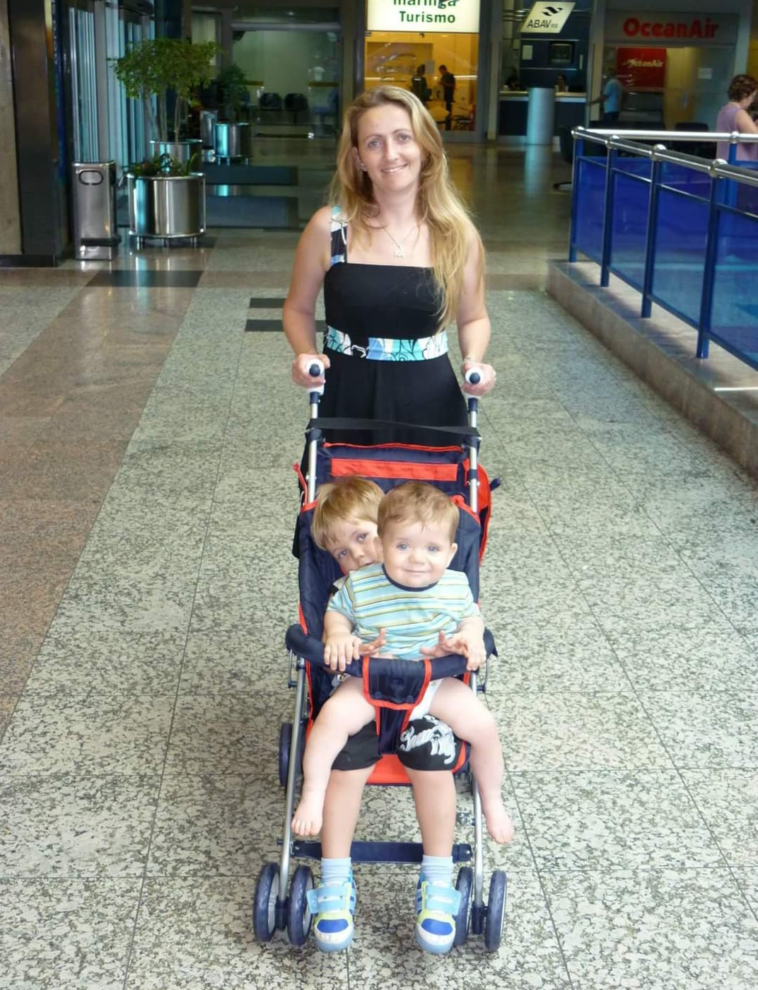Mulher empurrando um carrinho com duas crianças pequenas no saguão de um aeroporto.