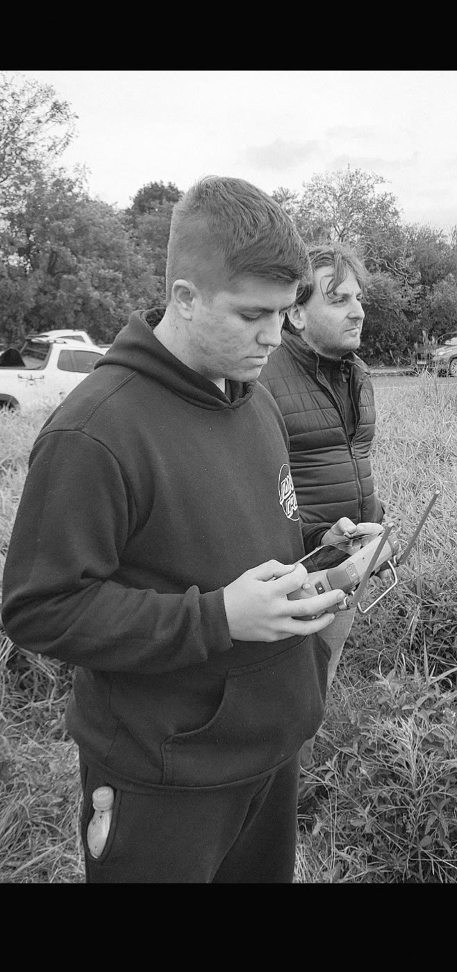 Foto em preto e branco de Johann no mato, olhando para o controle do drone, com outro homem atrás.